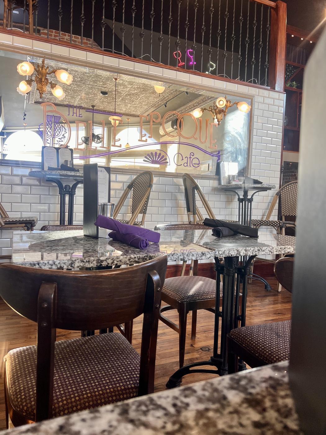 The dining room: a large mirror painted with the Belle Epoque wordmark in copper and purple, white subway tile, brass sconces and French rattan bistro chairs at speckled stone tables.